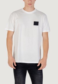 Camiseta de algodón blanca con cuello redondo y mangas cortas. Presenta un bolsillo rectangular negro en el lado izquierdo del pecho con detalle de logo en relieve.