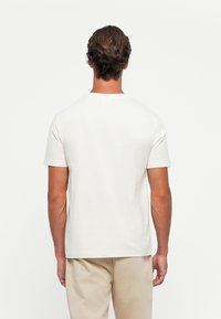 Mann mit kurzen braunen Haaren, der ein schlichtes weißes Kurzarm-T-Shirt und beigen Hosen trägt und mit dem Rücken zu einem weißen Hintergrund steht.