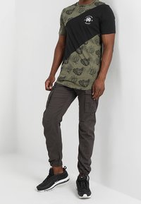 T-Shirt mit kurzen Ärmeln in einem Schwarz- und Olivgrün-Farbblockdesign mit Blattmustern, kombiniert mit dunklen Cargo-Hosen und schwarzen Sneakers.