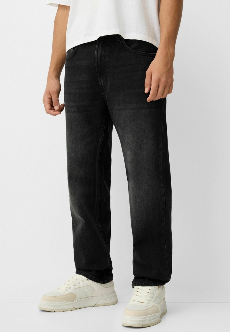 Vaqueros de mezclilla negra con un corte clásico, presentando una textura suave y un ligero desvanecimiento, combinados con zapatillas blancas.