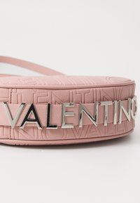 Rozā ādas soma ar reljefiem logotipa rakstiem un izteiksmīgu sudraba "VALENTINO" uzrakstu priekšpusē. Gluda tekstūra, taisnstūra forma.