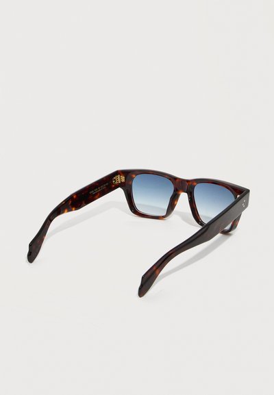 Γυαλιά ηλίου με σκελετό χελώνας και τετράγωνα μπλε φακούς με διαβάθμιση, εμφανιζόμενα από γωνιακή πίσω όψη σε λευκό φόντο.