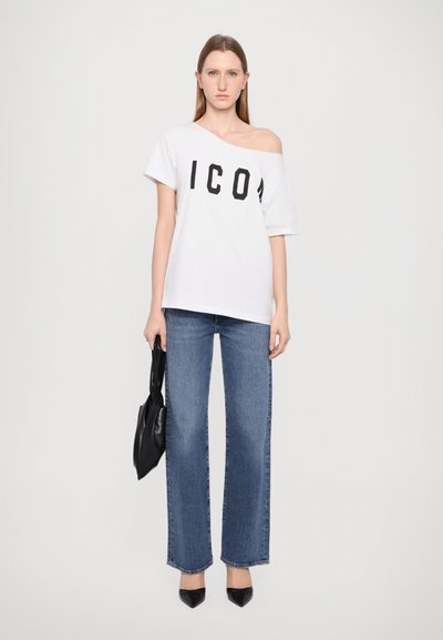 T-shirt blanc à manches courtes avec "ICON" en lettres noires, associé à un jean bleu évasé taille haute et des escarpins noirs à bout pointu. Sac noir à la main.
