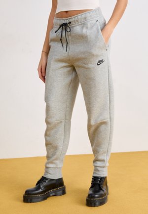 Osoba ubrana w szare spodnie dresowe Nike z czarnymi sznurkami i czarne masywne kozaki sznurowane, stojąca na żółto-brązowej podłodze.