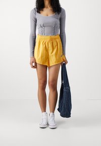 Grå långärmad topp med texturerade sömmar, matchad med ljusgula shorts med elastiskt midjeband och sidostråk. Vita sneakers och en denimväska.