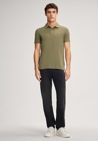 Olijfgroene polo met korte mouwen en een kraag, gecombineerd met donkerdenim broek en witte sneakers. Eenvoudig ontwerp met minimale branding.