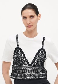 Čierny čipkový bralet cez biele tričko s krátkym rukávom, zdobené kvetinovými vzormi a zvlnenými okrajmi, s nastaviteľnými ramienkami a skráteným strihom.