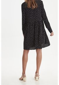 Abito lungo nero a maniche lunghe con pois bianchi, vita arricciata e gonna fluente. Indossato con sandali con tacco dorato, che mettono in mostra gambe scoperte.