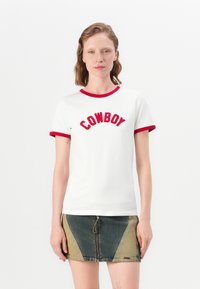 Valkoinen puuvillapaita, jossa on punainen kaula-aukko ja hihansuut, ja jossa on paksulla punaisella brodeerattu teksti "COWBOY". Yhdistetty farkkuhameeseen.