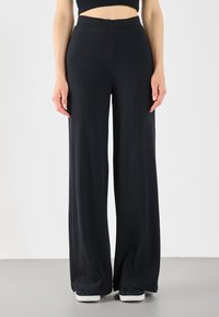 Pantalones de pierna ancha negros, hechos de material acanalado, con una cintura alta y un acabado texturizado. Llevados con zapatillas negras y blancas.