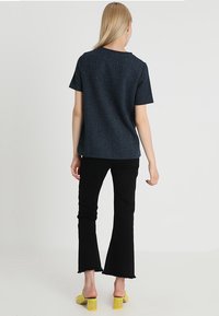 Mørkeblå top med korte ærmer i let struktureret stof; kombineret med sorte vide jeans og gule sandaler med blokhæl.