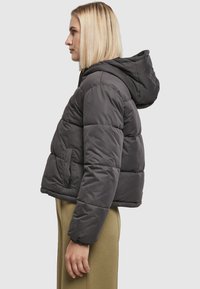 Vatteret jakke i mørkegrå, med hætte, quiltet mønster, sidelomme og elastiske manchetter. Materialet virker blødt og isolerende.