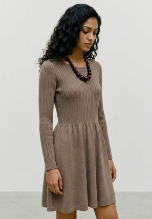 Femme aux longs cheveux bouclés portant une robe marron tricotée à manches longues et un collier foncé à perles, se tenant devant un fond clair uni.