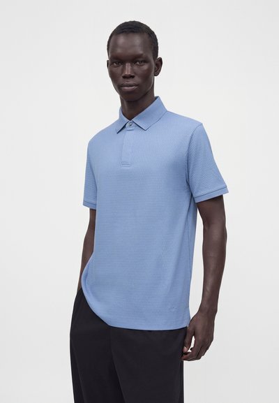 Homme portant un polo bleu clair texturé et un pantalon noir, debout devant un fond clair uni.