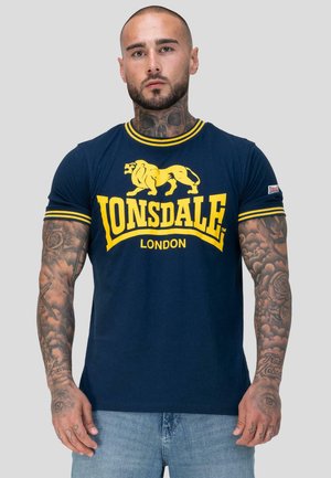 Mand med tatoveringer på nakke og arme iført en navy blå Lonsdale London t-shirt med gul logo og stribede ærmer, stående foran en grå baggrund.