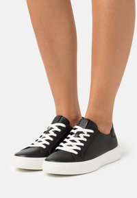 Sneaker in pelle nera con lacci bianchi, punta rotonda e suola in gomma bianca testurizzata. Presenta un logo sulla linguetta e una finitura liscia.