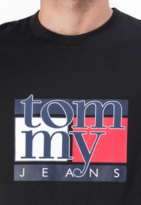 T-shirt noir avec un design graphique incluant "tommy" en bleu et "JEANS" en blanc, accentué par un fond rectangulaire rouge et bleu.