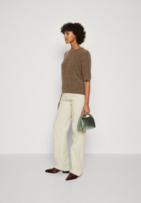 Maglione a coste marrone, pantaloni larghi crema e stivali borgogna a punta. Tiene una piccola borsa verde con manico superiore.
