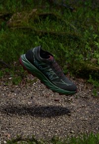Leichte Laufschuhe in Dunkelgrün mit Tarnmuster, roten Akzenten und gepolsterter Sohle, schwebend über einem Kiesweg.