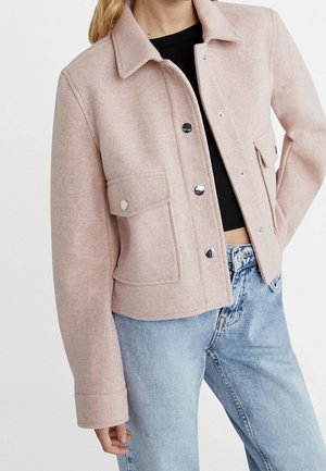 Woman wearing a light pink buttoned wool jacket with large front pockets over a black top and light blue jeans.