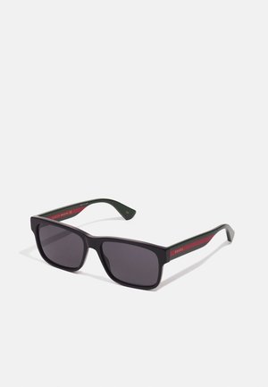 Gafas de sol rectangulares negras con lentes oscuros. Fabricadas en plástico, con brazos verdes que tienen una franja roja y acentos de logo en dorado.