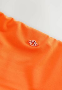 Felrood oranje stof heeft een gladde textuur met een klein zilveren logo accent nabij de bovenkant. Het materiaal is rekbaar en licht van gewicht.