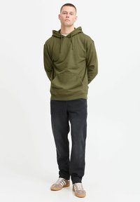 Olive green hooded sweatshirt with a front pocket, paired with dark jeans and brown sneakers featuring white stripes and a textured upper.