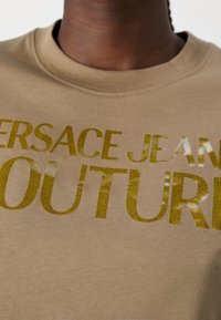 Beige katoenen t-shirt met vet, glinsterend gouden tekst die "VERSACE JEANS COUTURE" zegt. Ronde halsontwerp met korte mouwen.