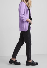 Kvinna som går, klädd i en lila kavaj, svart topp, svarta smala jeans och svarta kraftiga loafers mot en ljus, enkel bakgrund.