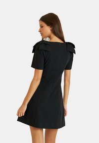 Schwarzes kurzärmliges Kleid mit Schleißdetails an den Schultern, tailliertem Oberteil und ausgestelltem Rock. Es verfügt über einen verdeckten Reißverschluss hinten zum Schließen.