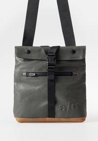 Gray waterproof bag with a fold-over top, black strap, zippered front pocket, and tan base; features logo embossed on the side.