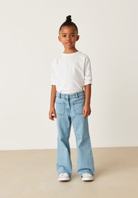 Cămașă albă cu mâneci lungi răsucite, asortată cu blugi evazați de denim albastru deschis, cu două buzunare la spate și adidași albi.