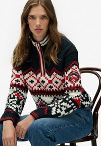 Gebreide trui met hoge kraag, marineblauw, rode en witte geometrische patronen, met geribde mouwen en een logo patch op de mouw.