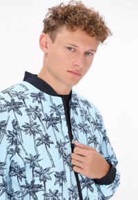 Giacca bomber azzurra chiara con stampa nera di palme, colletto e polsini a coste; presenta una zip frontale e una texture liscia.