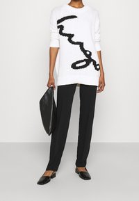 Maglione bianco lavorato a maglia oversize con design astratto nero, abbinato a pantaloni neri e una clutch nera, con scarpe nere flat.