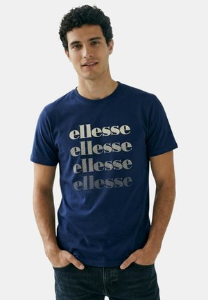 Homme aux cheveux bouclés et foncés portant un t-shirt bleu marine avec le mot "ellesse" imprimé quatre fois en blanc, debout les mains dans les poches.