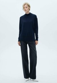 Pulover turtleneck bleumarin dintr-un material moale, asociat cu blugi largi de culoare închisă și pantofi cu vârf ascuțit albi. Design simplu și modern.
