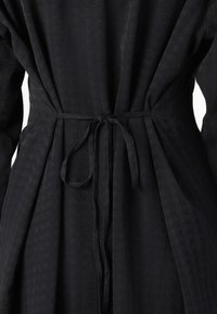 Vêtement noir en tissu texturé. Présente une taille cintrée à nouer, des manches longues et un léger plissé dans le dos. Design simple et élégant.