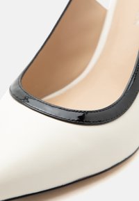 Gros plan sur une chaussure blanche pour femme avec une bordure noire brillante autour de l'ouverture et une doublure intérieure beige.