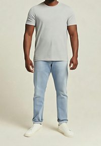 Homme debout portant un t-shirt uni gris clair à manches courtes, un jean bleu clair avec des revers, et des baskets blanches sur un fond neutre.
