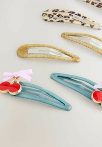 Hårnåle i forskellige designs: guld glimmer, leopardprint, og teal med et rødt hjerte og en lyserød sløjfe. Glatte og matte teksturer er synlige.