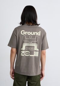 En person står med ryggen til beskueren, if�ørt en grå t-shirt med et stort grafisk design og tekst. Grønne bukser er synlige.