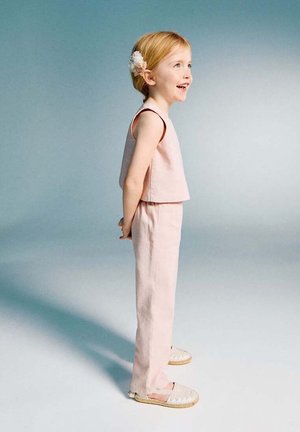 Fată tânără cu agrafă de flori în păr, purtând maiou fără mâneci de culoare roz deschis și pantaloni, zâmbind și uitându-se spre dreapta pe un fundal simplu.