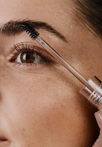 Brow Gel in einer klaren Tube mit einem schwarzen Pinsel, auf dunklen Augenbrauen aufgetragen. Nahaufnahme eines Auges mit subtilen Sommersprossen und glatter Hautstruktur.