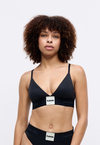Juodas Calvin Klein braletas su V formos iškirpte, plonomis petnešėlėmis ir kontrastiniu logotipu apatinėje dalyje. Lygus audinys, minimalistinis dizainas.