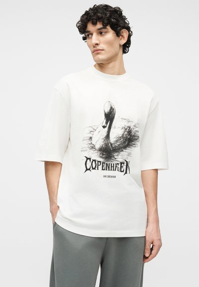 Νέος άνδρας με σγουρά μαλλιά, φορώντας μια λευκή υπερμεγέθη μπλούζα με μαύρο σχέδιο κύκνου και την λέξη "Κοπεγχάγη", συνδυασμένη με γκρι παντελόνι.