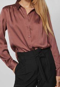 Satäng, vinröd blus med lång ärm, knappframsida och rynkad axeldetalj, kombinerad med högmidjade svarta byxor med knytband.