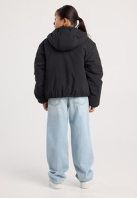 Young person with ponytail wearing a black hooded puffer jacket, light blue wide-leg jeans, and white sneakers, facing away from the camera.