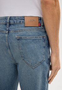 Zbliżenie na osobę noszącą niebieskie dżinsy z brązową skórzaną metką Joop! Jeans umieszczoną nad prawą tylną kieszenią oraz czerwoną metką na pasku talii.