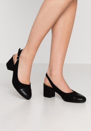 Nogi w czarnych sandałach na blokowym obcasie typu slingback, zaokrąglonym noskiem i srebrną klamrą po boku, stojące na białej podłodze.
